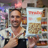 A smiling man in a shop holds up a bag of Trafo Sweet Popped Corn organic snack. The background shows shelves stocked with various snacks and treats, with a pink neon 'Treats' sign hanging above.