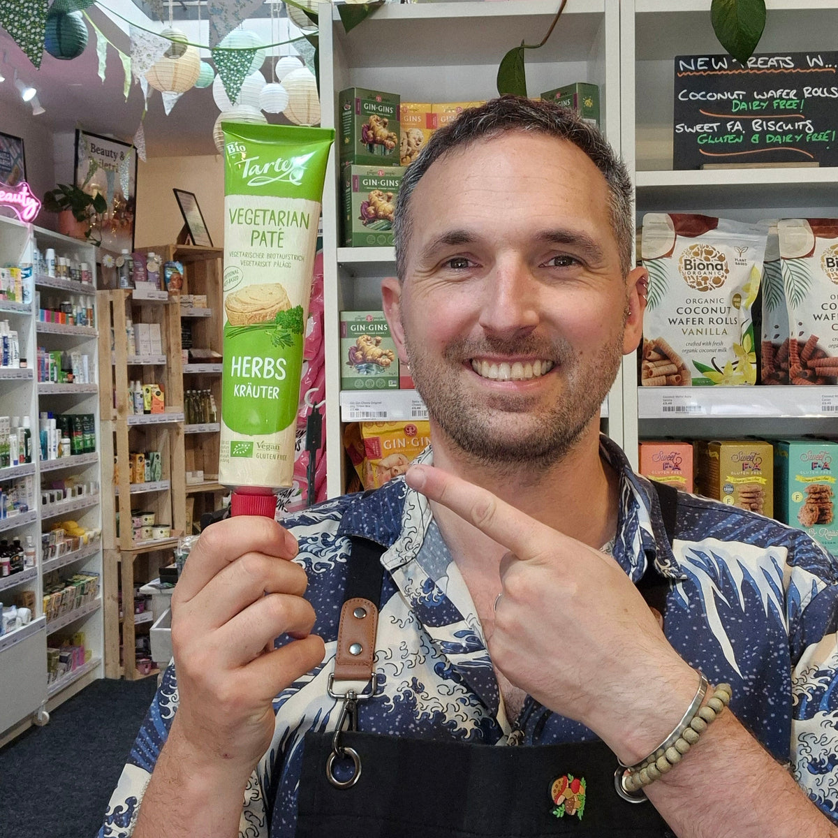A smiling man wearing a black Health & Remedy apron over a blue and white patterned shirt, posing in a retail store with shelves of products in the background.