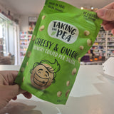 A person holds a green bag of Taking the Pea Cheesy & Onion Crunchy Coated Pea Snack in a shop setting.