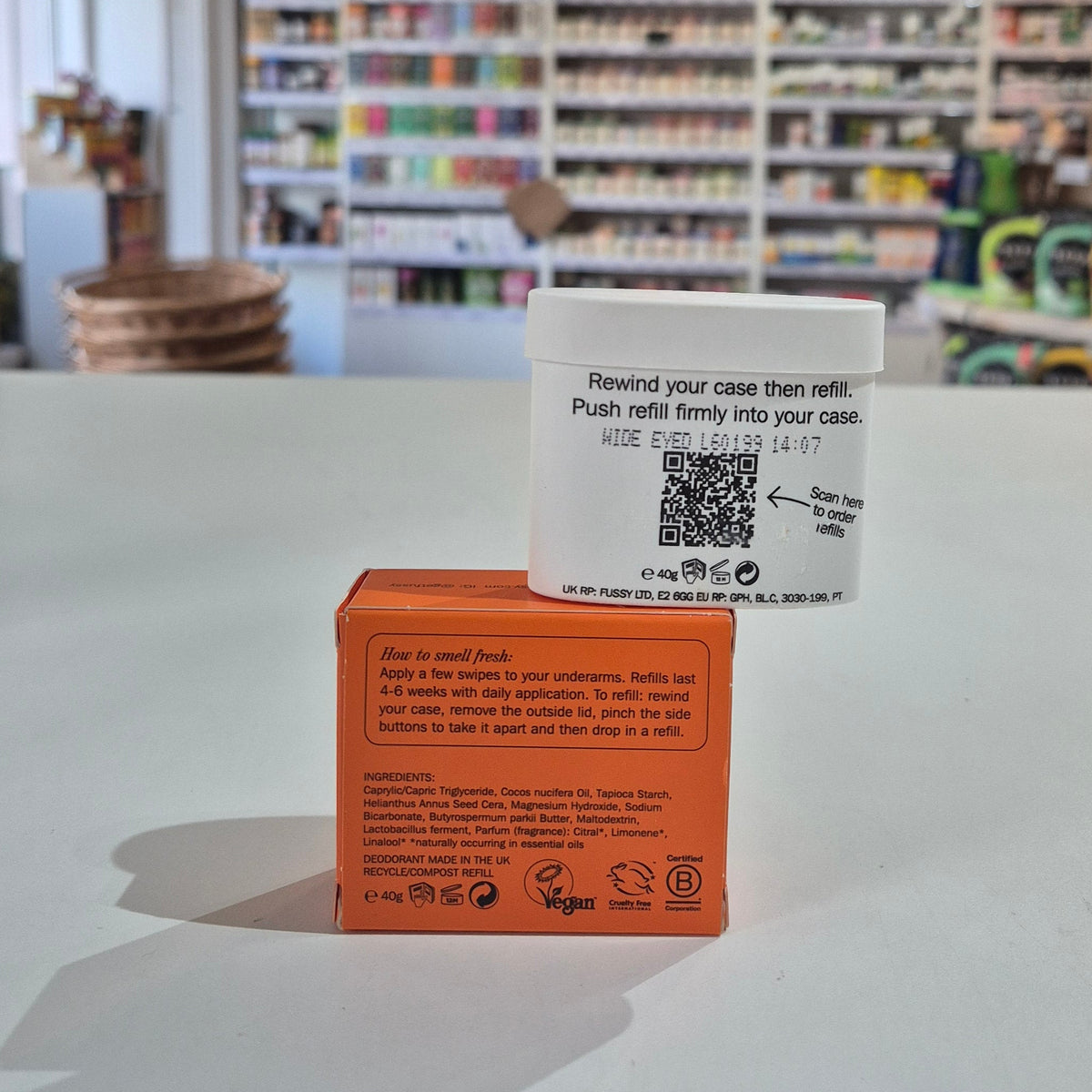 Fussy compostable deodorant refill container and packaging on a table with a blurred store background. White container with a QR code label on a red box in a store setting