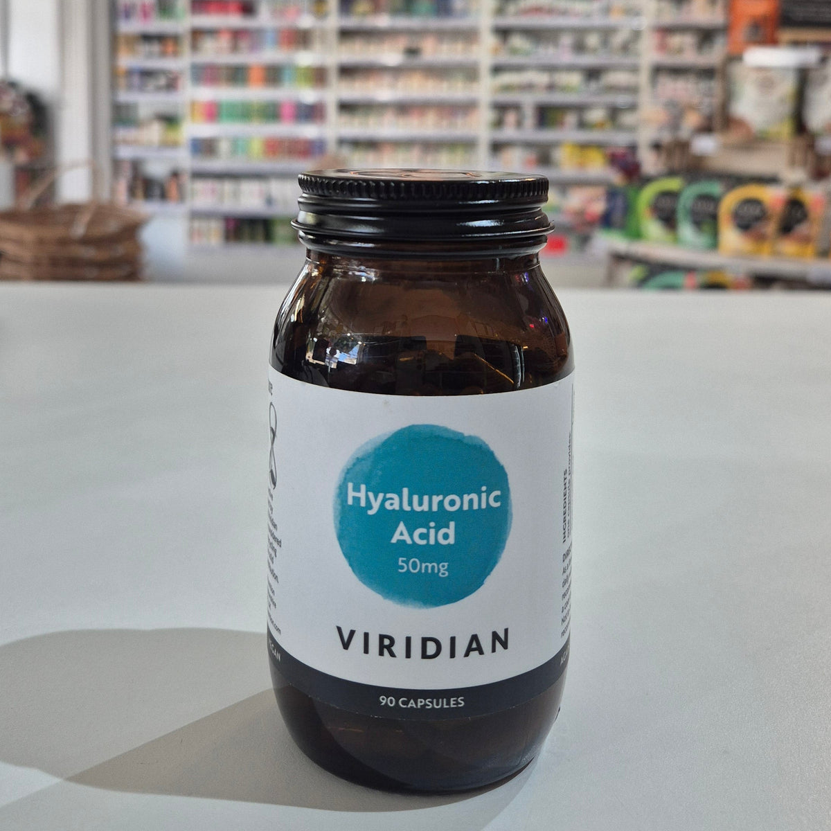 Brown supplement bottle labeled 'Hyaluronic Acid' by Viridian on a white surface with a blurred store background.