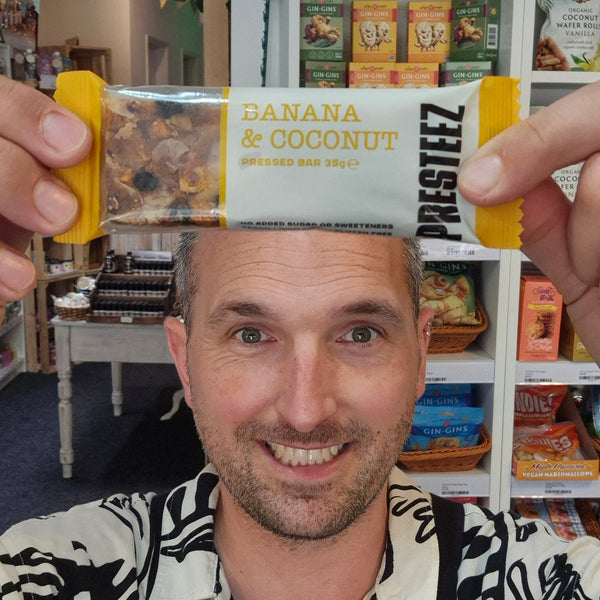 A smiling man in a patterned shirt holds a bar of Prestez Banana & Coconut spread in front of his face in a retail shop setting.