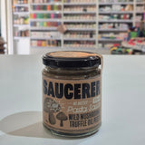 A glass jar of Saucerer Wild Mushroom Truffle Oil pesto with a black lid and brown label, sitting on a white table in a blurred shop interior.