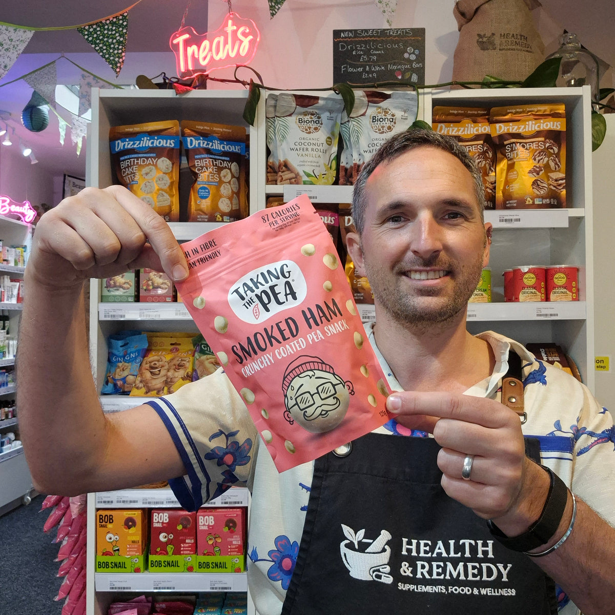 A man wearing a black Health & Remedy apron holds up a pink pouch of 'Baking Pea Smoked Ham' in a retail store setting with shelves of snacks behind him.