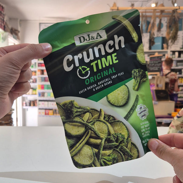 A person holds a bag of Dj&A Crunch Time Original vegetable chips in a store setting. The bag is green and black with a photo of green vegetable chips on the front.