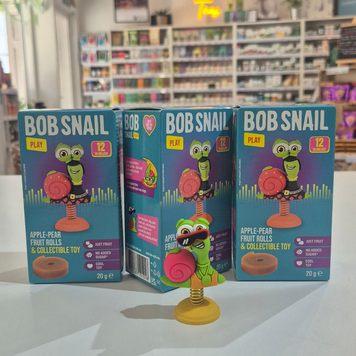 Three Bob Snail fruit roll and collectible toy packages on a table with a store shelf in the background.