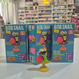 Three Bob Snail fruit roll and collectible toy packages on a table with a store shelf in the background.