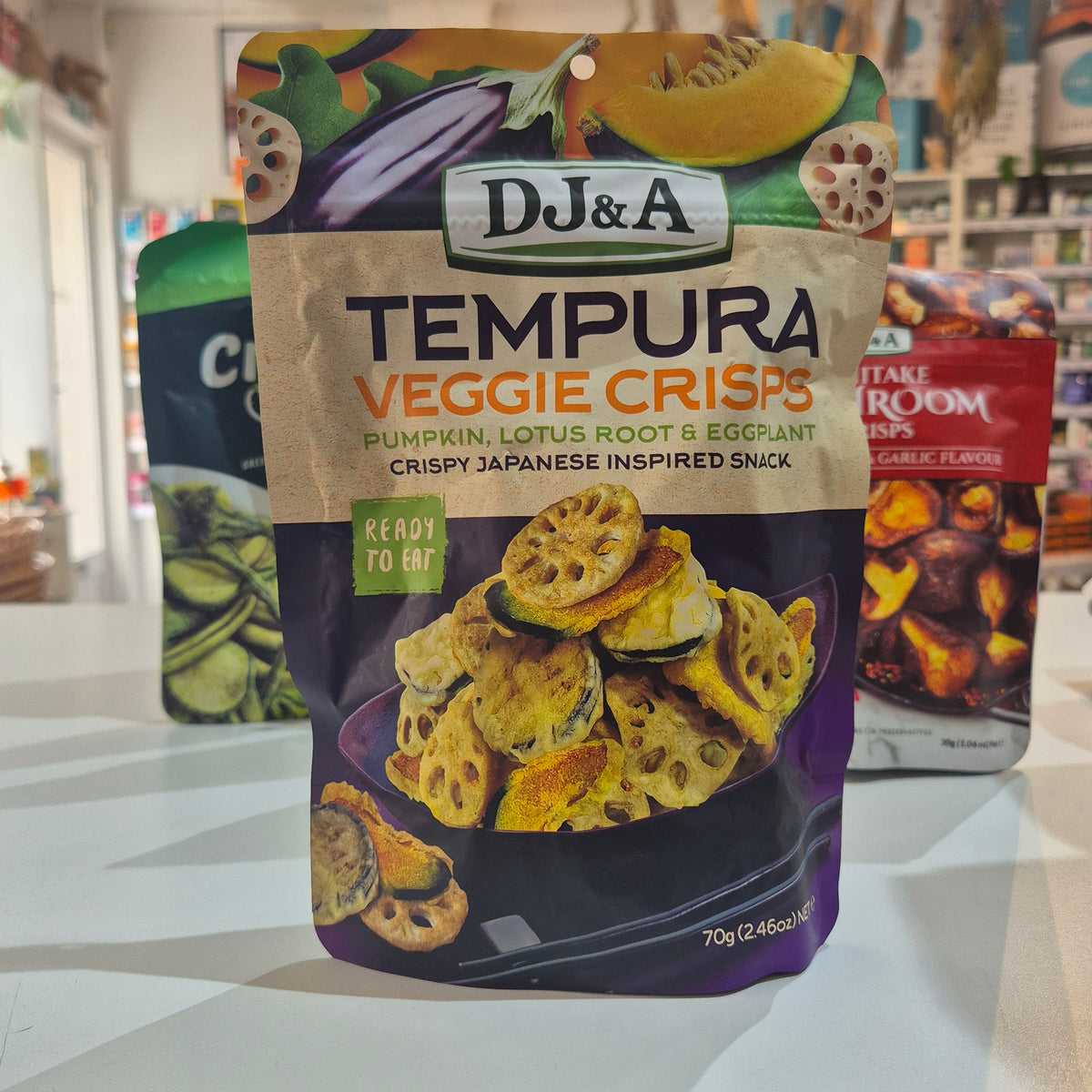 A bag of DJ&A Tempura Veggie Crisps (pumpkin, lotus root, and eggplant flavors) standing on a white counter in a store. The packaging features a kraft paper style top half with colorful illustrations of vegetables, and a dark purple bottom half showing the crispy snack pieces.
