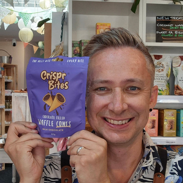 A smiling man in a kitchen or cafe setting holds up a purple bag of Crisper Bites Waffle Cones. He is wearing a black apron with a white logo and a colourful patterned shirt.
