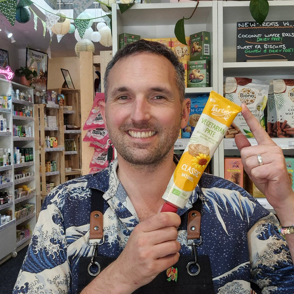 A smiling man in a blue and white patterned shirt and black apron holds up a tube of Tartex Classic Pate in a brightly lit health shop.
