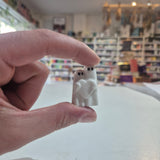 A hand holding a small white ceramic ghost figurine with black eyes and a heart-shaped cutout in the center, set against a blurred background of a shop interior.