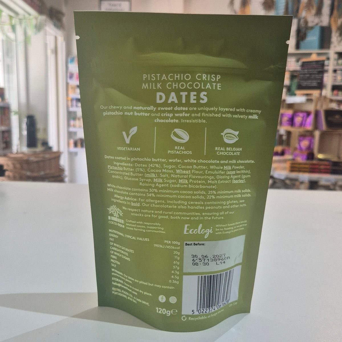 Green package of pistachio crisp milk chocolate dates on a white surface with a blurred store background.