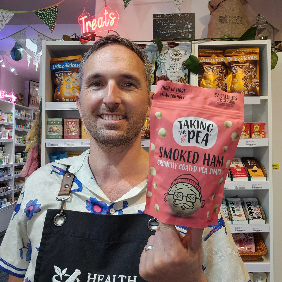 A man wearing a floral shirt and a black Health & Remedy apron stands in a retail shop holding a pink bag of 'Taking the Pea Smoked Ham Crunchy Coated Pea Snacks'.