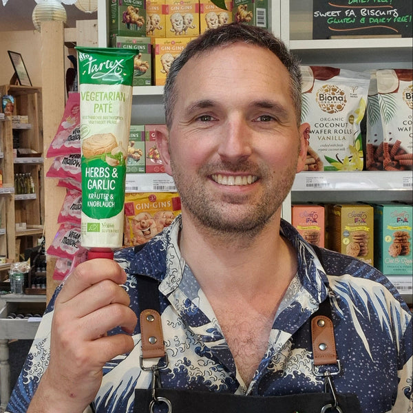 A man wearing a black Health & Remedy apron and a blue patterned shirt smiles while holding a Tartex tube in a retail store setting with shelves of products in the background.