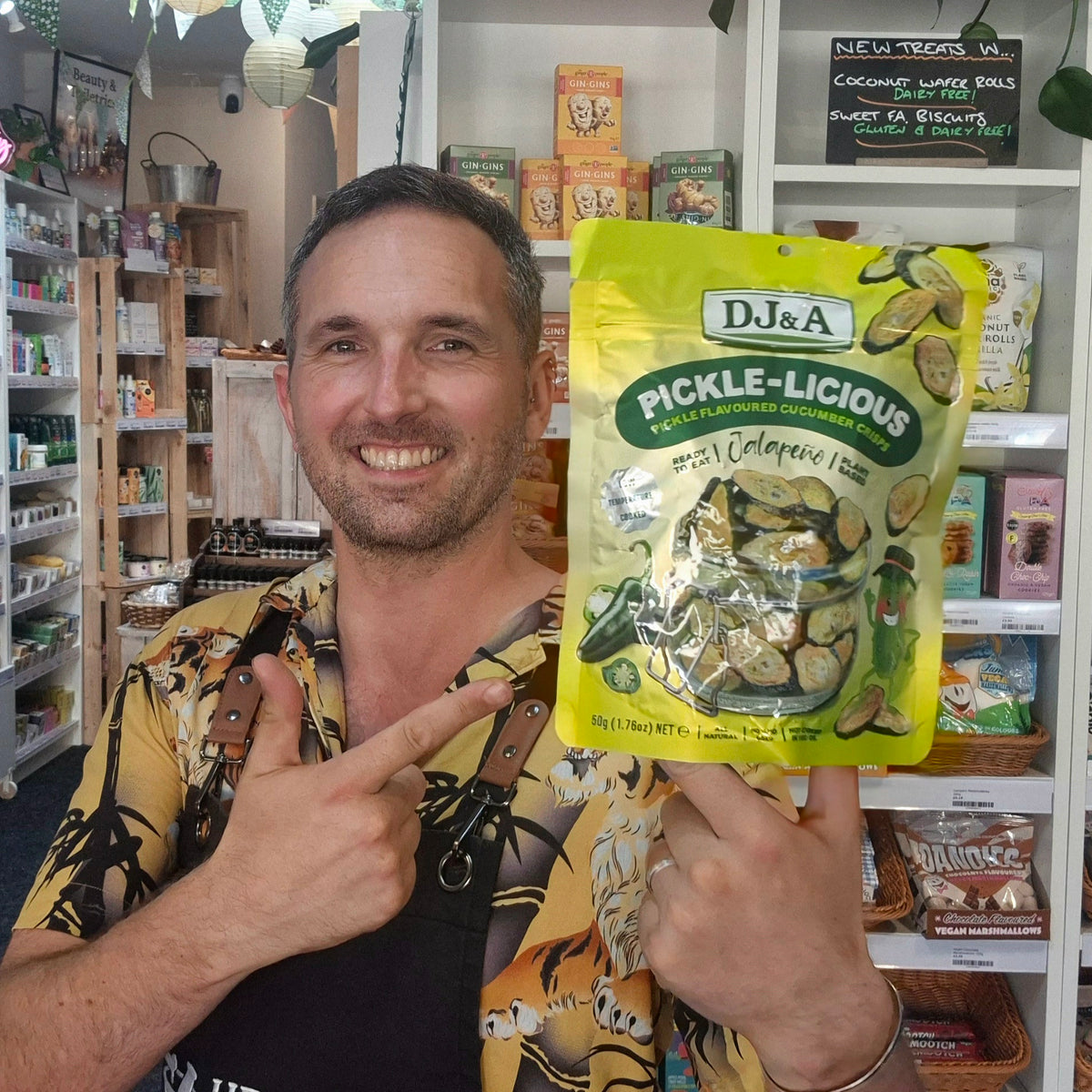 A smiling man in a yellow patterned shirt holds up a bag of DJ&A Pickle-Licious Pickled Jalapeno in a shop setting with shelves of goods in the background.