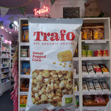 A hand holds a bag of Trafo Sweet Popped Corn organic snacks in a retail shop setting with shelves of other products in the background.