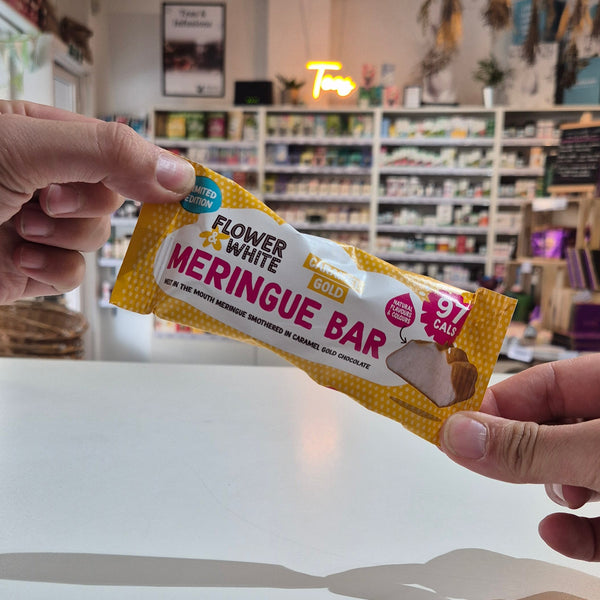 A hand holding a yellow and white packaged meringue bar, with another hand pointing at it. The background shows a blurred indoor shop setting with shelves and a white wall.