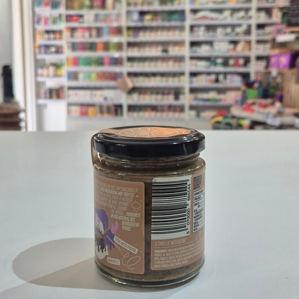 A glass jar of Saucerer Wild Mushroom Truffle Oil pesto with a black lid and brown label, sitting on a white table in a blurred shop interior.