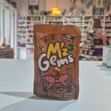 A bag of M'z Gems Novelty Nuts Milk Chocolate novelty chocolate shells, standing on a white counter in a blurred shop setting.