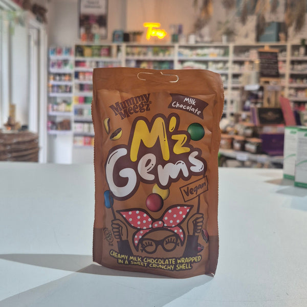 A bag of M'z Gems Novelty Nuts Milk Chocolate novelty chocolate shells, standing on a white counter in a blurred shop setting.