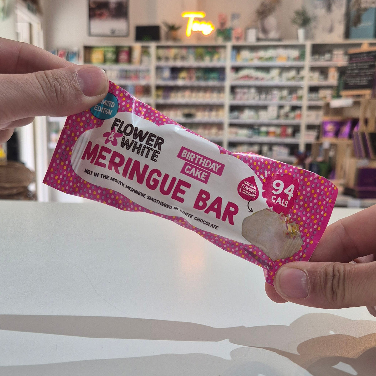 A person holds a packaged flower & white Birthday Cake Meringue Bar in a brightly lit shop setting. The packaging is pink with white polka dots, a white center, and red text.