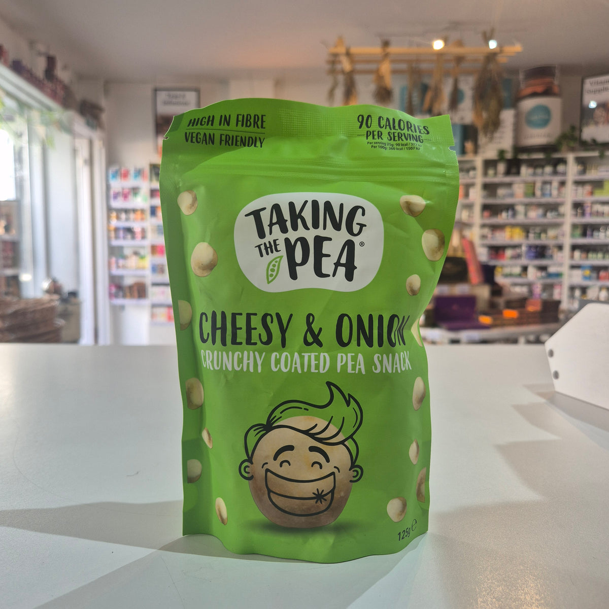 A green bag of Taking the Pea Cheesy & Onion Crunchy Coated Pea Snack standing on a white counter in a store setting with blurred shelves in the background.