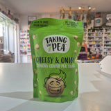 A green bag of Taking the Pea Cheesy & Onion Crunchy Coated Pea Snack standing on a white counter in a store setting with blurred shelves in the background.