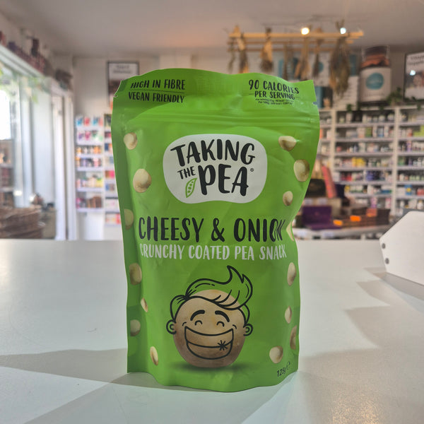 A green bag of Taking the Pea Cheesy & Onion Crunchy Coated Pea Snack standing on a white counter in a store setting with blurred shelves in the background.
