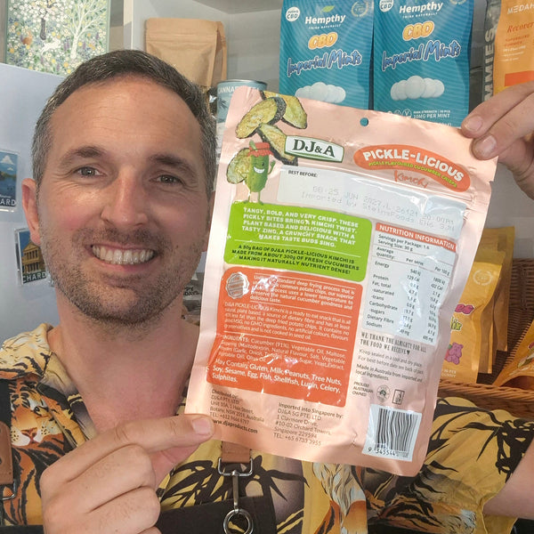 A smiling man in a tiger-print shirt holds up the back of a bag of D&J A Pickle-Licious Kimchi seasoning mix in a retail setting.