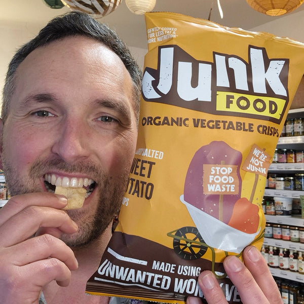 Man eating a snack from a 'Junk Food' organic vegetable crisps package in a store.