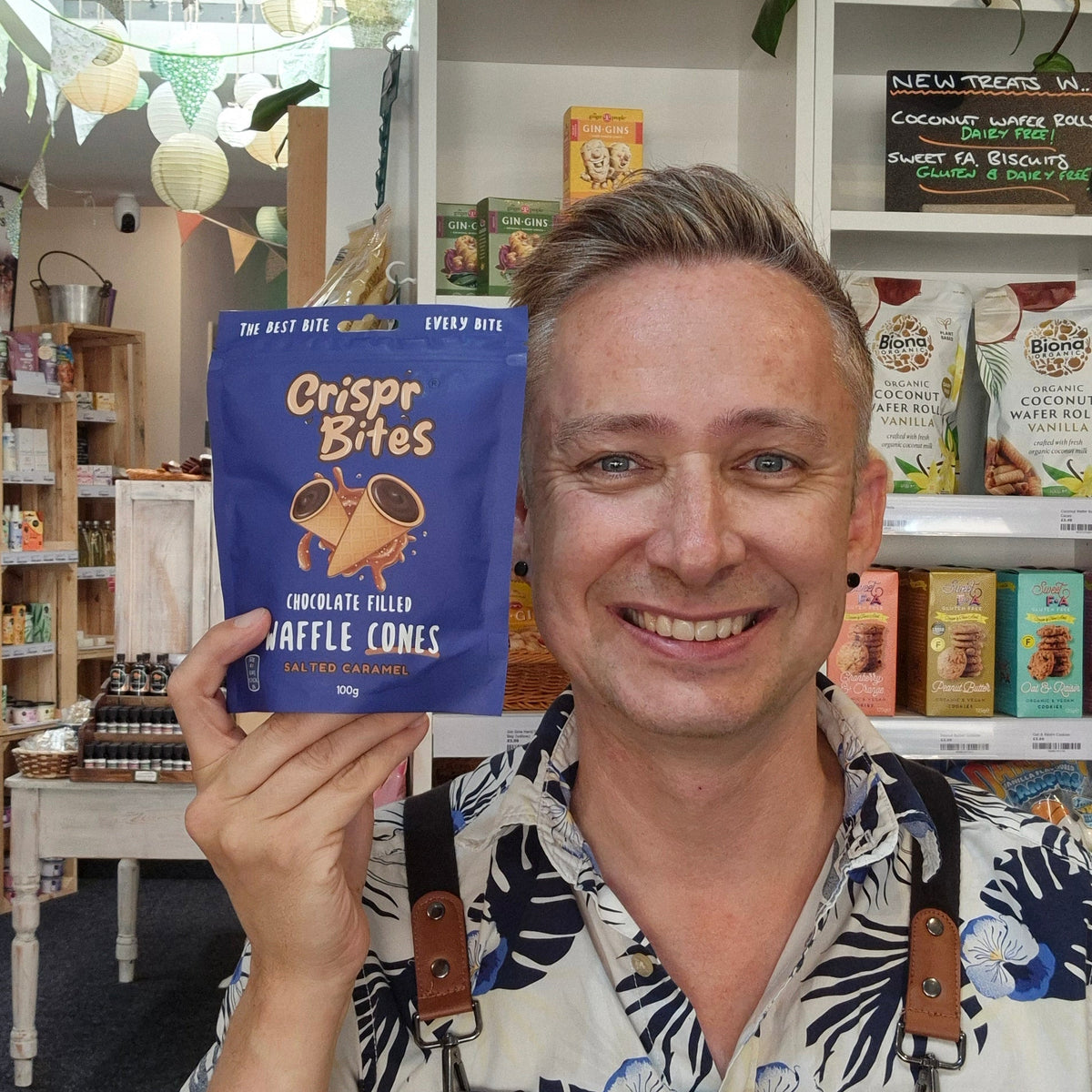 A smiling man in a floral shirt and Health & Remedy apron holds up a blue bag of Crisp Bites chocolate filled truffle cones in a shop setting.