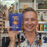 A smiling man in a floral shirt and Health & Remedy apron holds up a blue bag of Crisp Bites chocolate filled truffle cones in a shop setting.