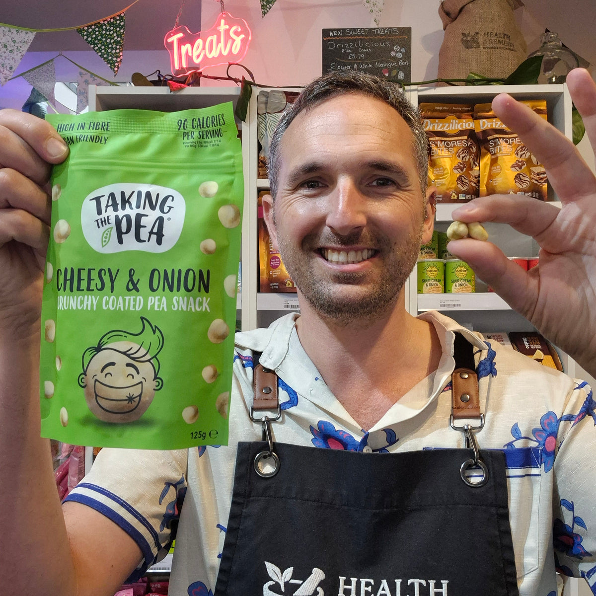 A smiling man in a kitchen or snack shop setting holds up a green bag of Taking the Pea Cheesy & Onion crunchy coated pea snack. He is wearing a black apron with the white logo 'Health & Remedy' and has shelves of packaged snacks behind him.