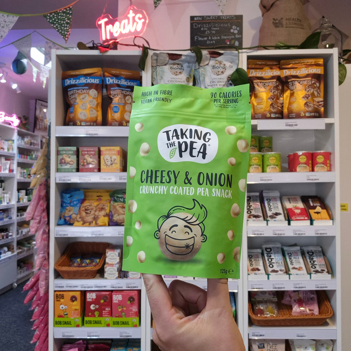 A hand holds a green bag of Taking the Pea 'Cheesy & Onion' crunchy coated pea snack in a retail store setting with shelves of other snacks behind it.