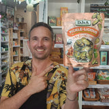 A man in a tropical print shirt and black apron holds up a bag of DJ&A Pickle-Licious Pickled Kimchi n a retail shop setting.