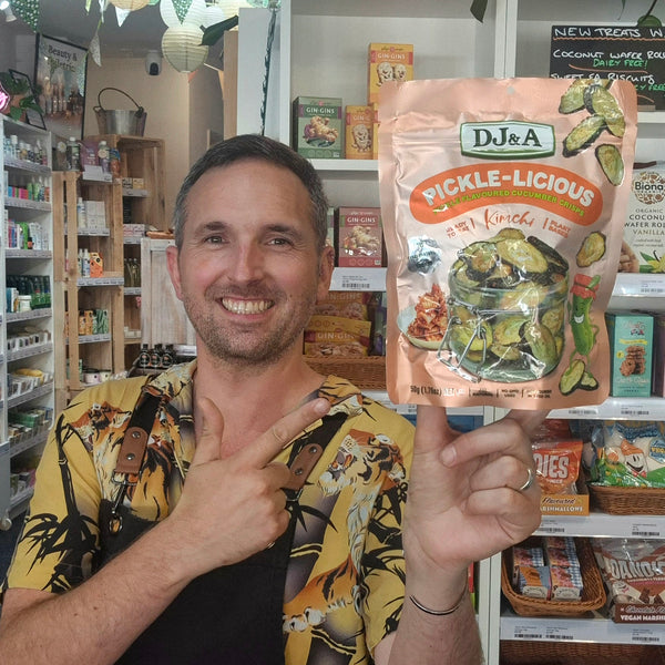 A man in a tropical print shirt and black apron holds up a bag of DJ&A Pickle-Licious Pickled Kimchi n a retail shop setting.