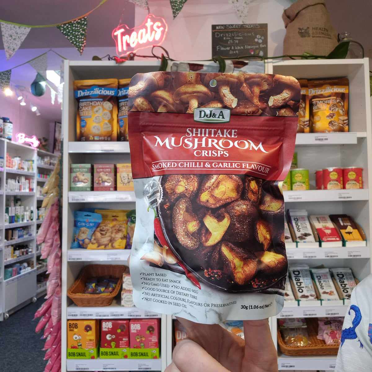 A hand holds a bag of Dj&A Shitake Mushroom Crisps in the smoked chili & garlic flavour, positioned in front of a retail store shelf filled with other snack products.