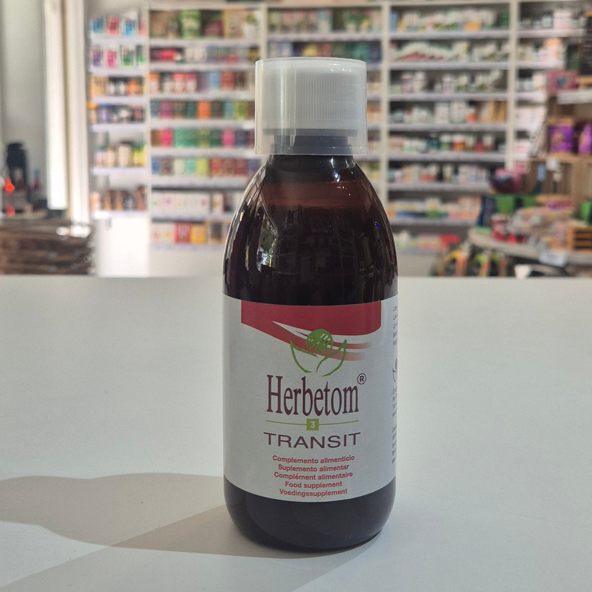 A bottle of Herbetom Transit food supplement with a white cap and white label, set on a white surface with a blurred pharmacy background.