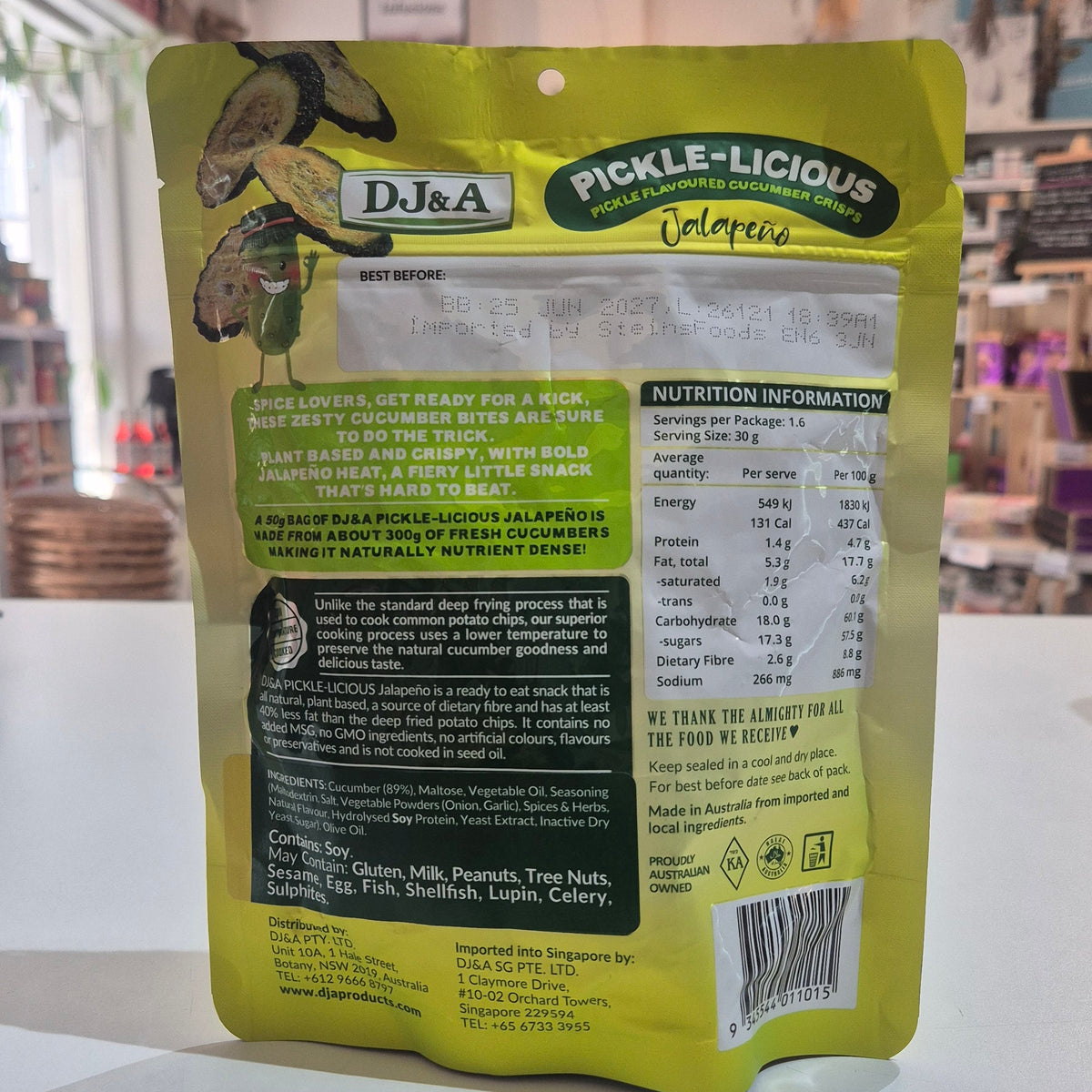 A yellow and green stand-up pouch of D&J&A Pickle-Licious showing the back of the package with ingredients and nutrition facts, set on a white surface in a shop.