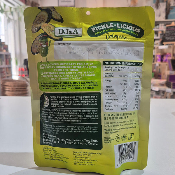 A yellow and green stand-up pouch of D&J&A Pickle-Licious showing the back of the package with ingredients and nutrition facts, set on a white surface in a shop.