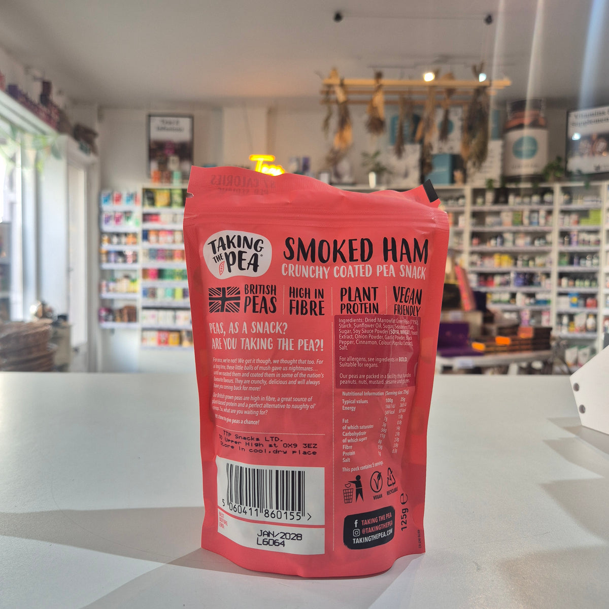 A red stand-up pouch of Taking 'Pea Smoked Ham Crunchy Coated Pea Snacks, photographed from the back on a white tabletop with a blurred shop interior in the background.