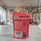 A red stand-up pouch of Taking 'Pea Smoked Ham Crunchy Coated Pea Snacks, photographed from the back on a white tabletop with a blurred shop interior in the background.