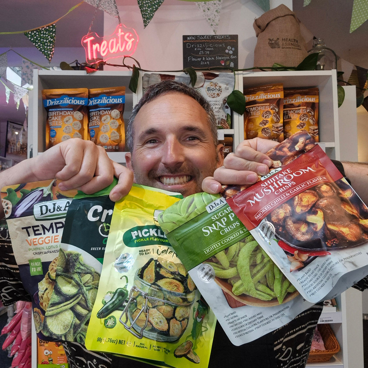 A smiling man holds up four bags of gourmet snacks and vegetables in front of a store shelf. The bags include DJ&A