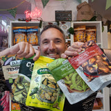 A smiling man holds up four bags of gourmet snacks and vegetables in front of a store shelf. The bags include DJ&A