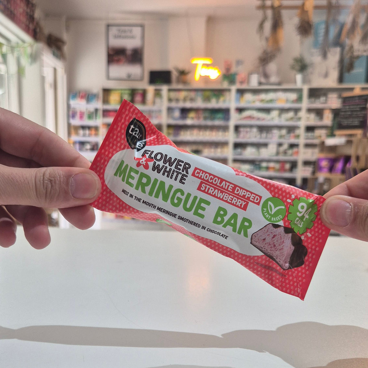 A person's hands holding a packaged meringue bar in front of a blurred shop background. The package is red with white and green text reading 'Flower White Chocolate Dipped Strawberry Meringue Bar'.