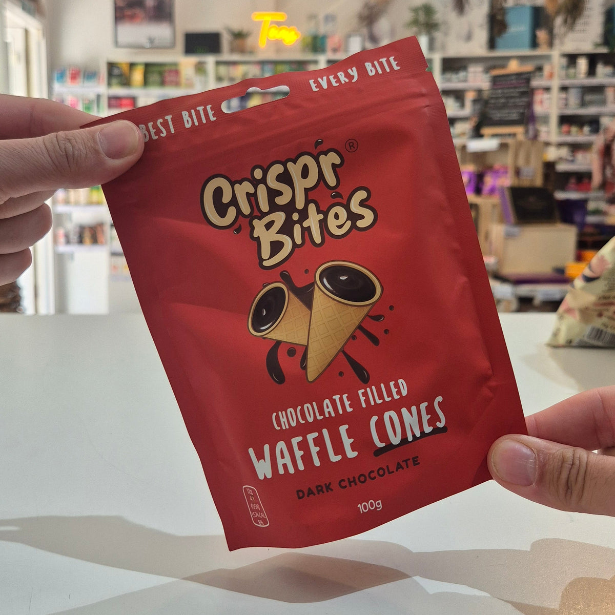 A person holds a red bag of Crispr Bites Dark Chocolate Filled Waffle Cones in a brightly lit shop interior.