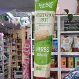 A hand holding a tube of Tartex vegetarian pâté with herbs flavour, set against a blurry background of a retail store shelf.