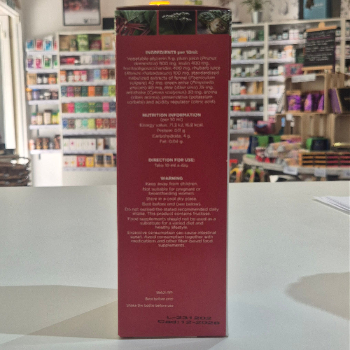 A tall red product box for Herbetom standing upright on a white surface, photographed in a retail store with shelves of other products blurred in the background. The front of the box is not visible, so only the back panel is shown, with small white text including ingredients, nutrition info, and directions.