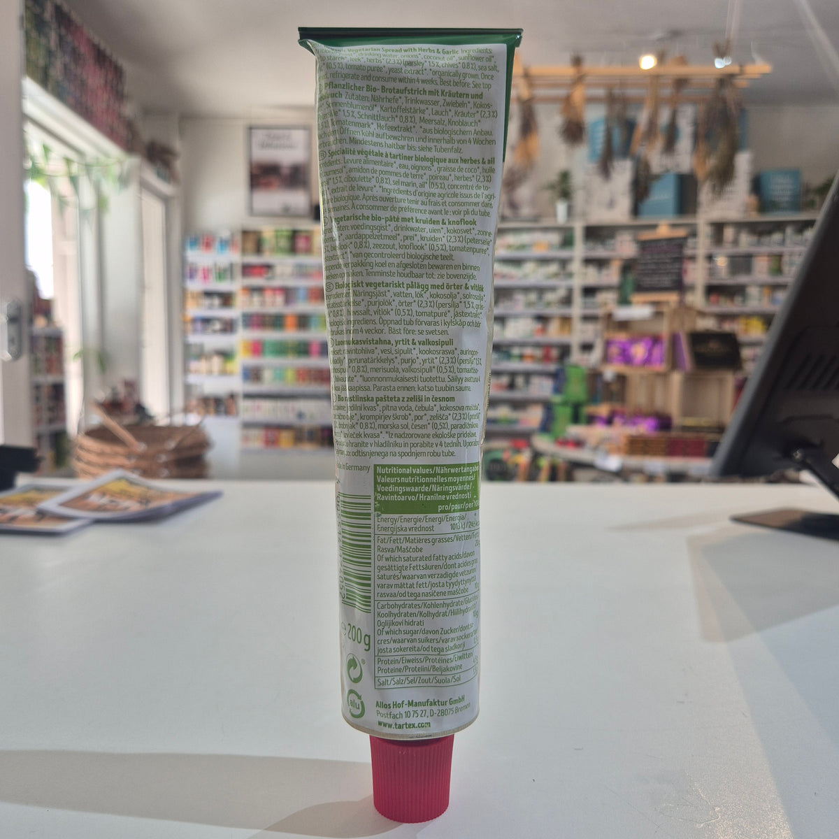 A squeeze tube Tartex Herbs and Garlic with a pink cap, lying horizontally on a white surface with a blurred store interior in the background.