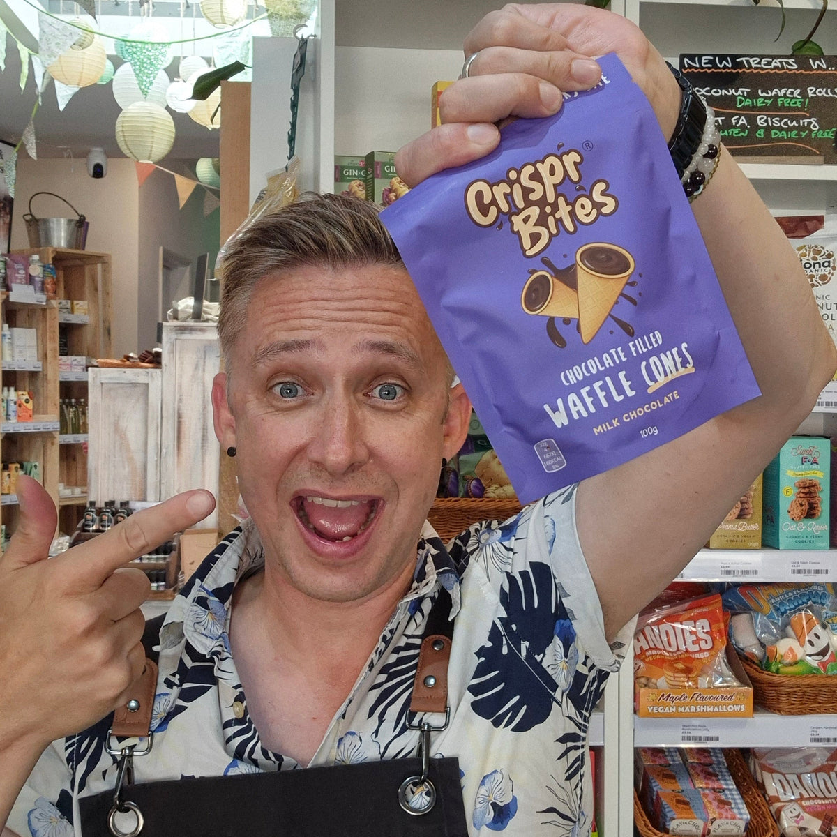 A man wearing a 'Health & Remedy' apron holds up a purple bag of 'crispr Bites' waffle chips in a retail shop setting.